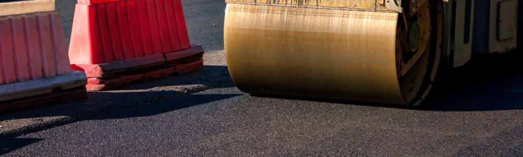 Workers are engaged in asphalt paving on a city road, utilizing heavy machinery to ensure a smooth surface. Bright sun illuminates the area, revealing safety barriers and ongoing construction efforts.