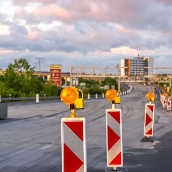 A line of road barriers with flashing lights during highway repair with greenery and urban background.